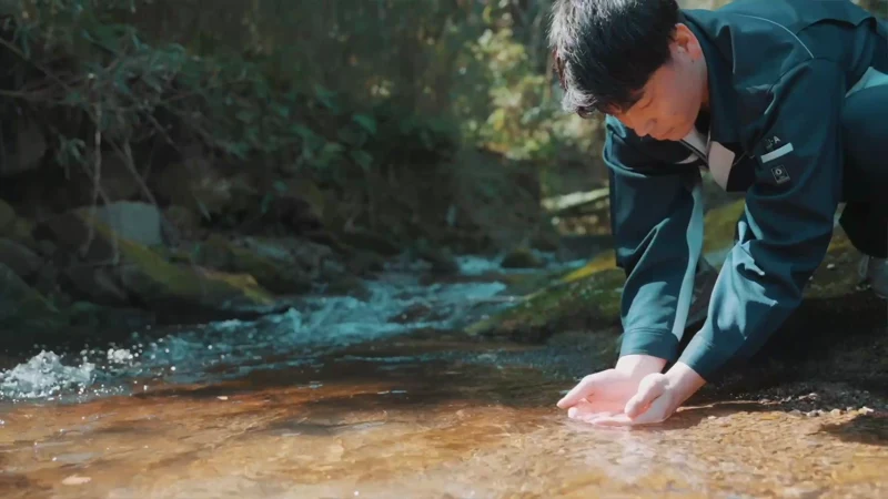 自然に囲まれた川辺で、しゃがんだスタッフが両手を水に浸している。